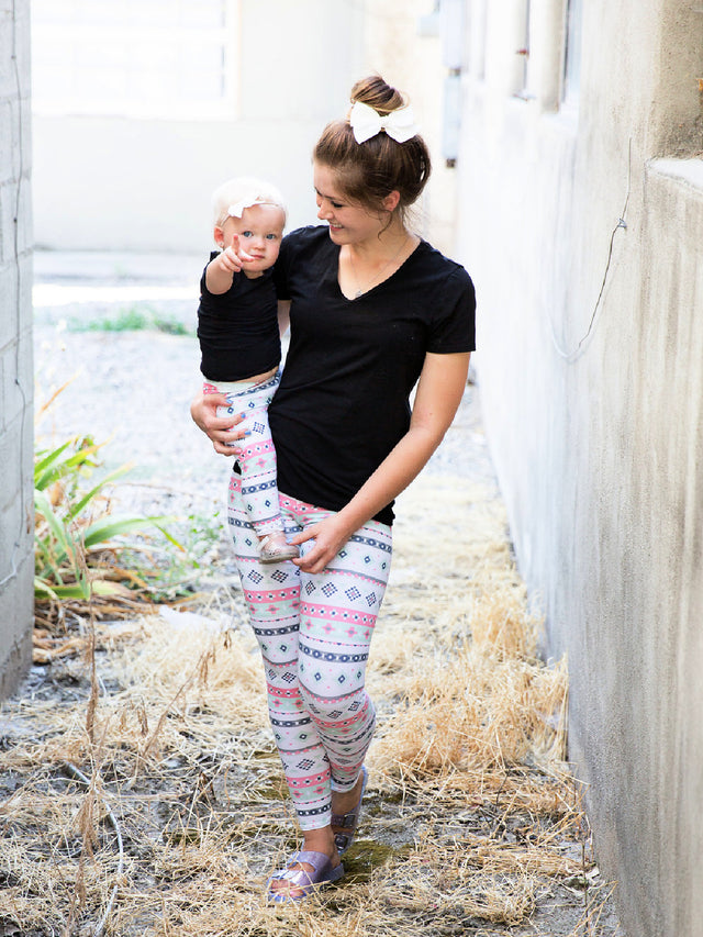 Woman wearing pastel aztec print leggings with matching baby, both in black tops and white bows, smiling outdoors.