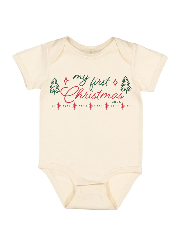 A cream-colored short-sleeve baby bodysuit featuring a festive graphic on the chest that reads 'my first Christmas 2026' in green and red script, flanked by small green pine trees and a dotted line with red stars.