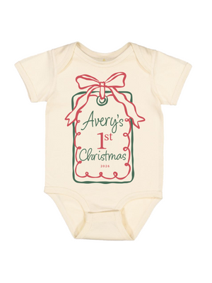 A cream-colored short-sleeve baby bodysuit featuring a personalized Christmas gift tag graphic on the front. The graphic has a pink bow, green border, and the text 'Avery's 1st Christmas 2026' in green and red lettering.