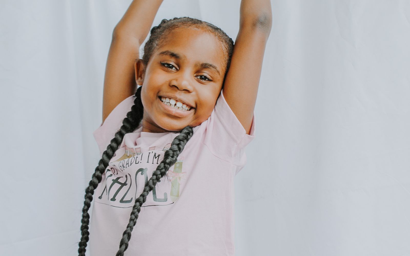 Smiling Black girl with long braids wearing a light pink personalized back-to-school shirt that says “Hello 2nd Grade! I’m Khloe” while raising her arms excitedly.