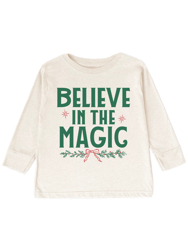 An off-white long-sleeve baby t-shirt with a festive graphic that reads 'BELIEVE IN THE MAGIC' in green, decorated with small red stars and a red bow on a green holly branch, shown on a plain white background.