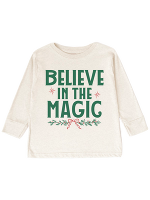 An off-white long-sleeve baby t-shirt with a festive graphic that reads 'BELIEVE IN THE MAGIC' in green, decorated with small red stars and a red bow on a green holly branch, shown on a plain white background.