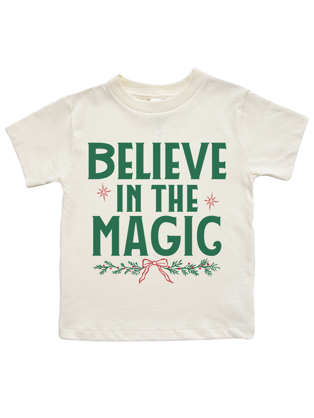 An off-white short-sleeve kids' t-shirt featuring a green graphic print that reads 'BELIEVE IN THE MAGIC' with small red starbursts, green pine branches, and a red bow on a white background.