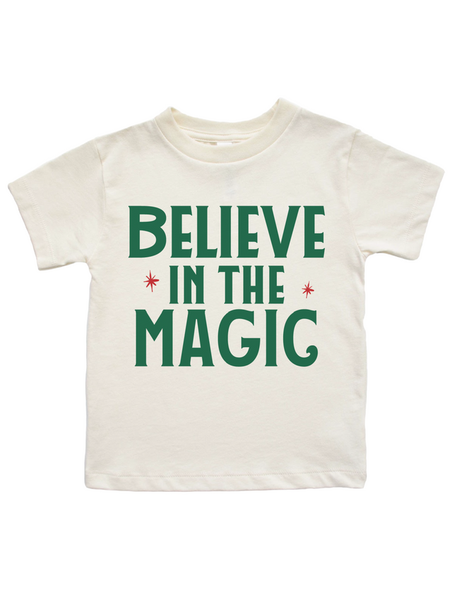 An off-white short-sleeve t-shirt with the phrase 'BELIEVE IN THE MAGIC' printed in dark green block letters, accented with small red starbursts, shown against a plain white background.