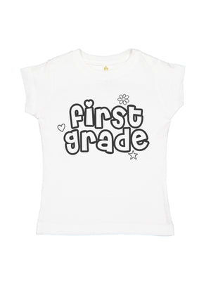 A white short-sleeve t-shirt for kids, featuring a black graphic print on the front that reads 'first grade' in a bubbly hand-drawn font, decorated with small heart, flower, and star outlines.