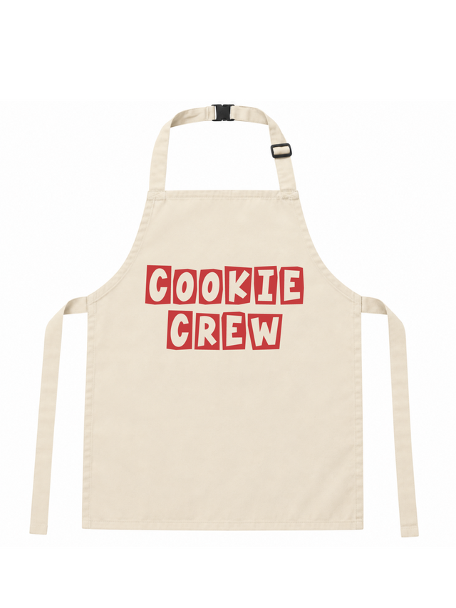 An off-white canvas apron with 'COOKIE CREW' printed in red block letters with white outlines across the chest, featuring a neck strap and waist ties.