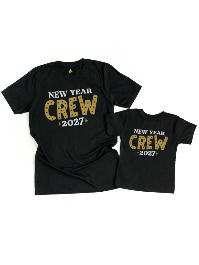 Two black crewneck t-shirts, one adult size and one child size, displayed flat against a white background. Both shirts feature a graphic print that reads 'NEW YEAR CREW 2027' in white and gold glitter-textured lettering.