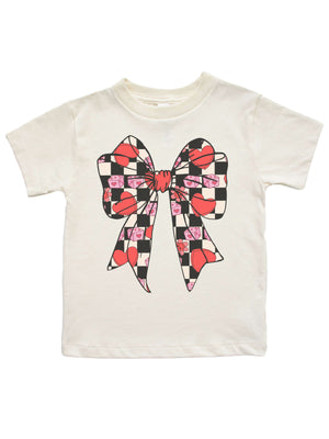 Natural-colored girls Valentine shirt featuring a retro bow design with checkered hearts, pink dice, and flowers, displayed flat on a white background.