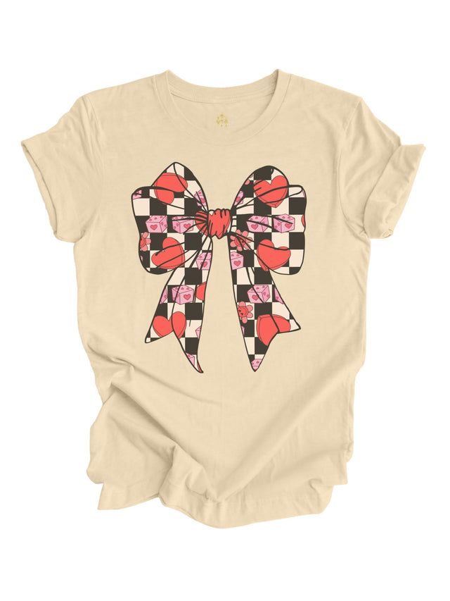 Natural-colored Coquette Valentine Bow shirt featuring a retro bow design with checkered hearts, pink dice, and flowers, displayed flat with rolled sleeves on a white background.