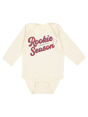 A cream-colored long-sleeve baby bodysuit featuring 'Rookie Season' printed in red script across the chest, accompanied by a small baseball graphic.