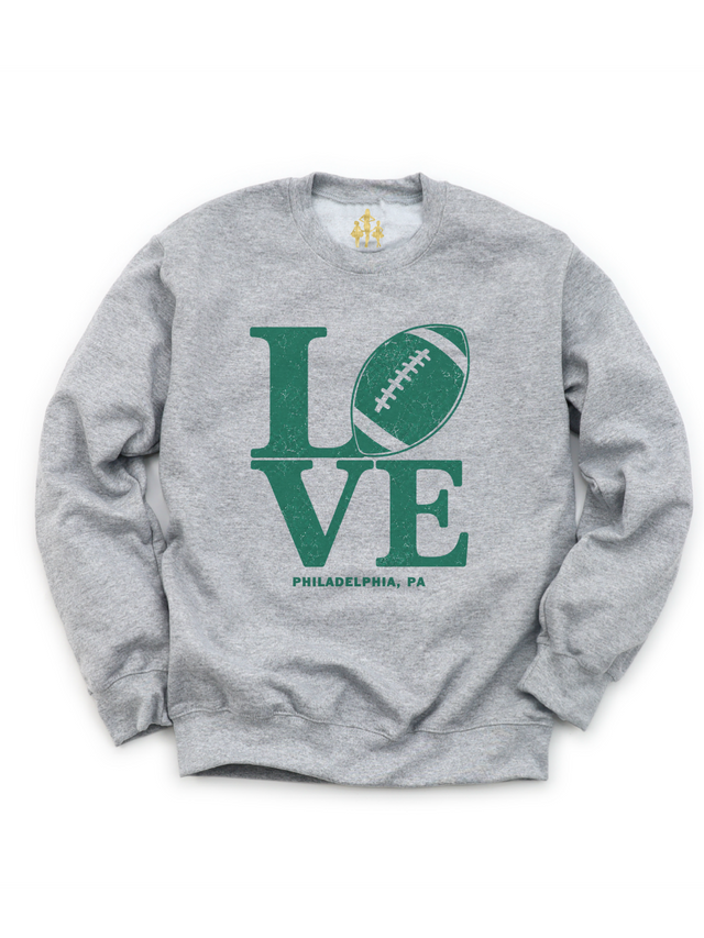 A heather gray crewneck sweatshirt featuring a green graphic that spells 'LOVE' with a football replacing the letter 'O', and 'PHILADELPHIA, PA' printed below in small green text.