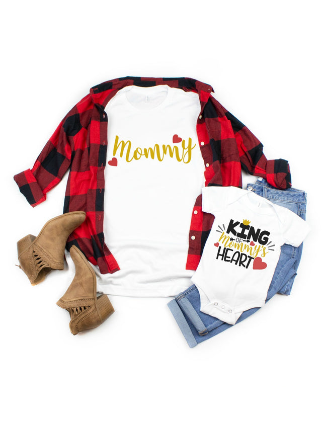 Mother and son matching Valentine's Day shirts. Women's white short sleeve shirt that says Mommy in gold with buffalo plaid jacket and fringe boots. Infant white bodysuit that says King of Mommy's heart on top of denim jeans