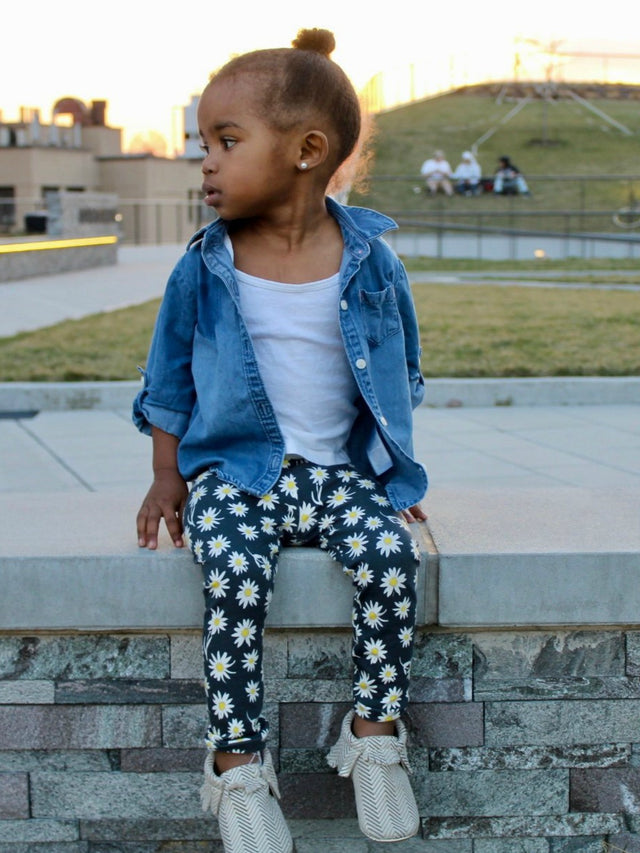 Toddler girl wearing black daisy print leggings, denim shirt, and white moccasins, sitting outdoors on a stone ledge at sunset.